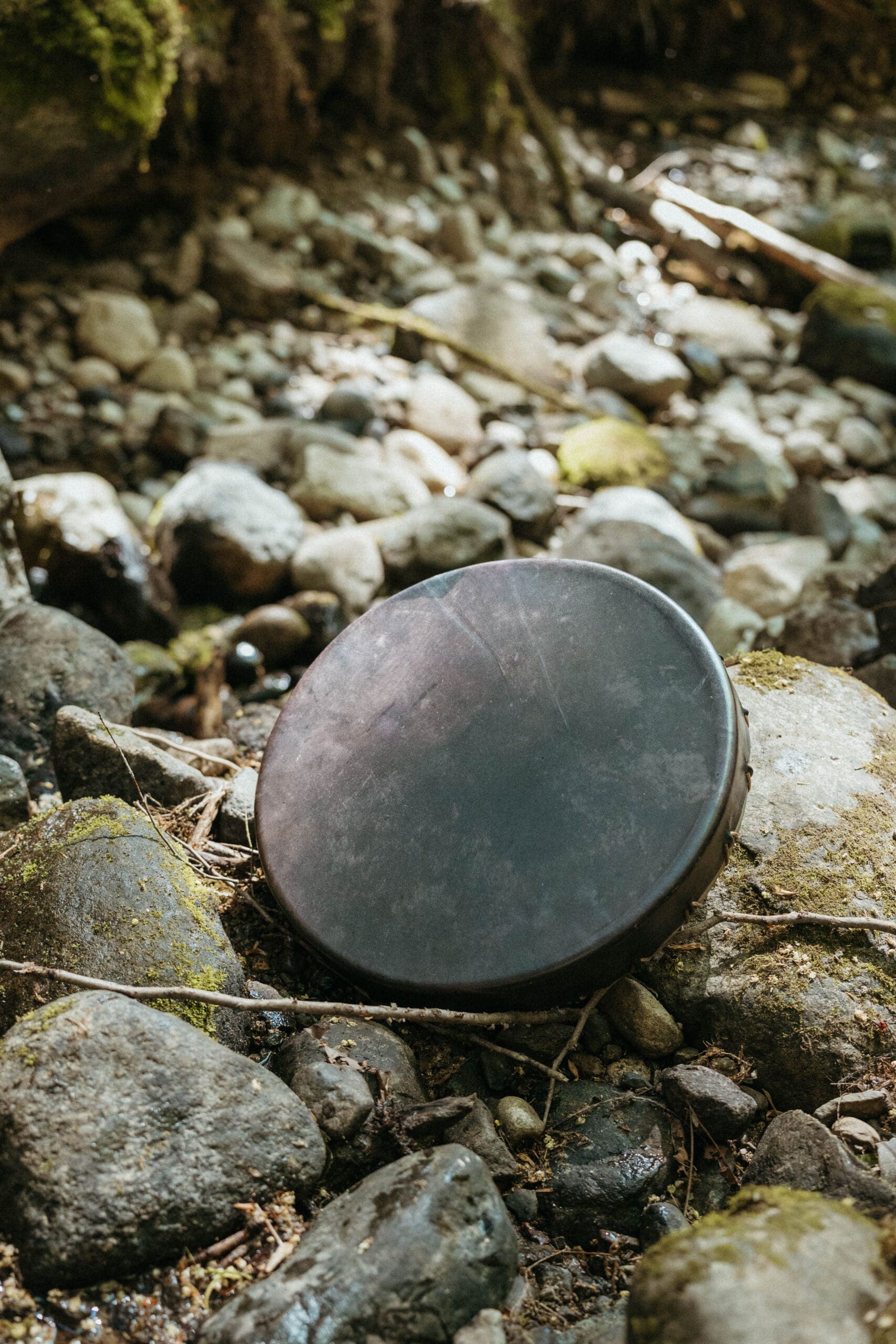 Black hand drum resting on river rocks, representing grounding, place, and beginning a gathering with care.