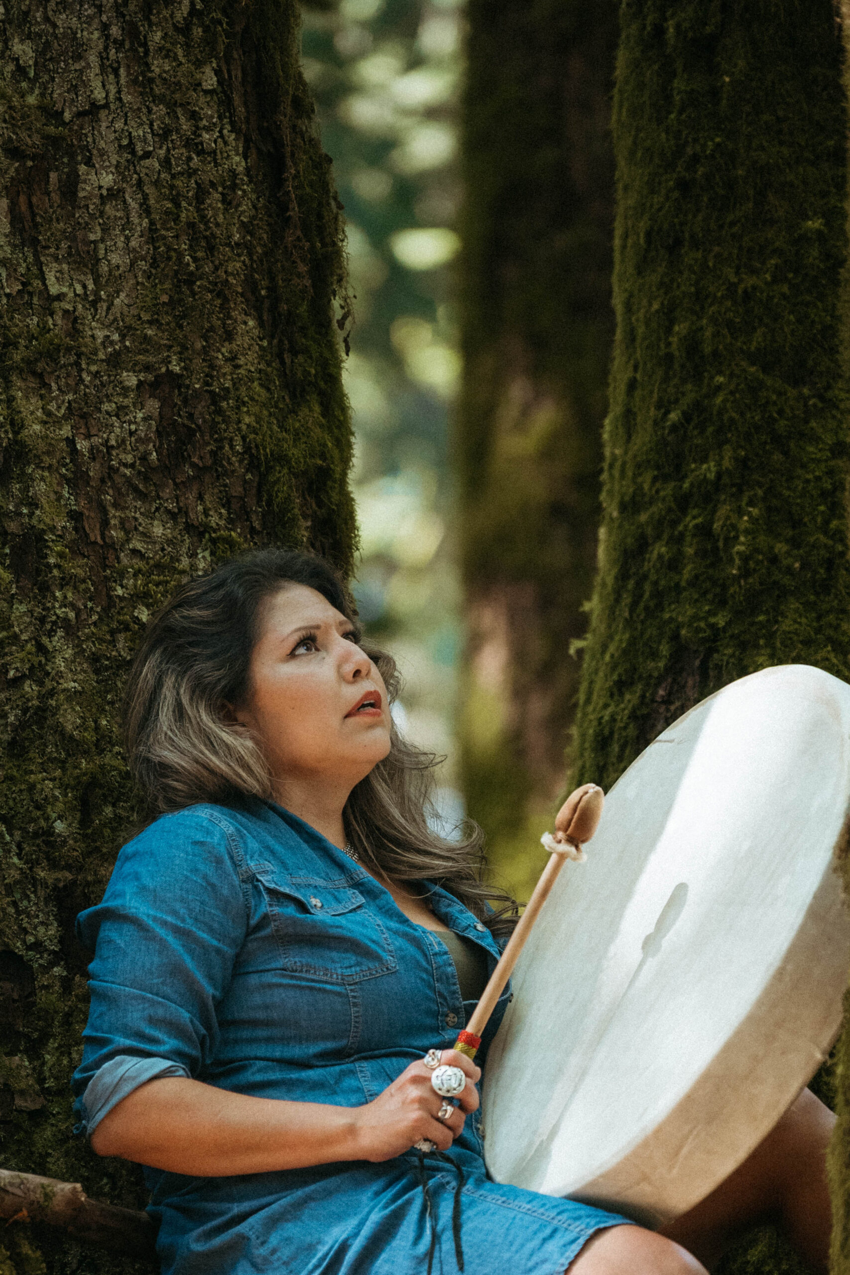 Naomi holding a hand drum between cedar trees